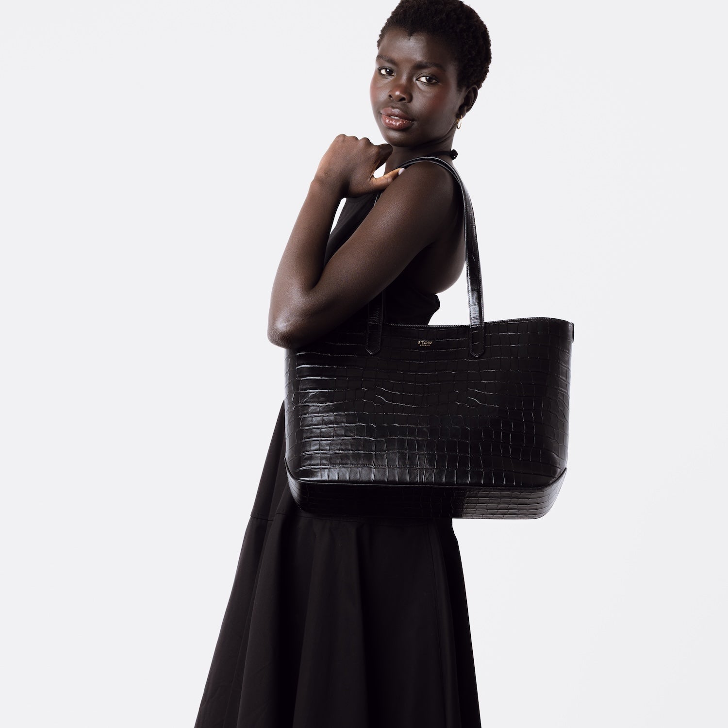 Model wearing Stow London's Everyday Tote Bag in Black Embossed Croc leather on her shoulder on a white background featuring gold STOW logo