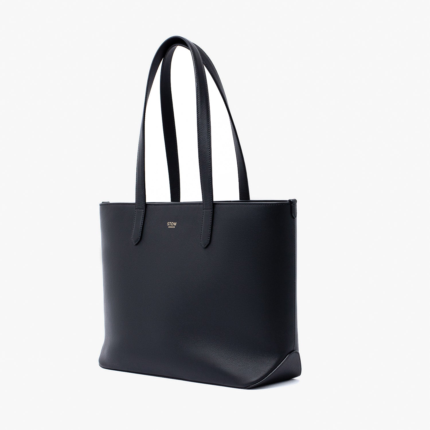 Off-axis view of Stow London's Everyday Tote Bag in Black smooth leather on a light grey background featuring gold STOW logo