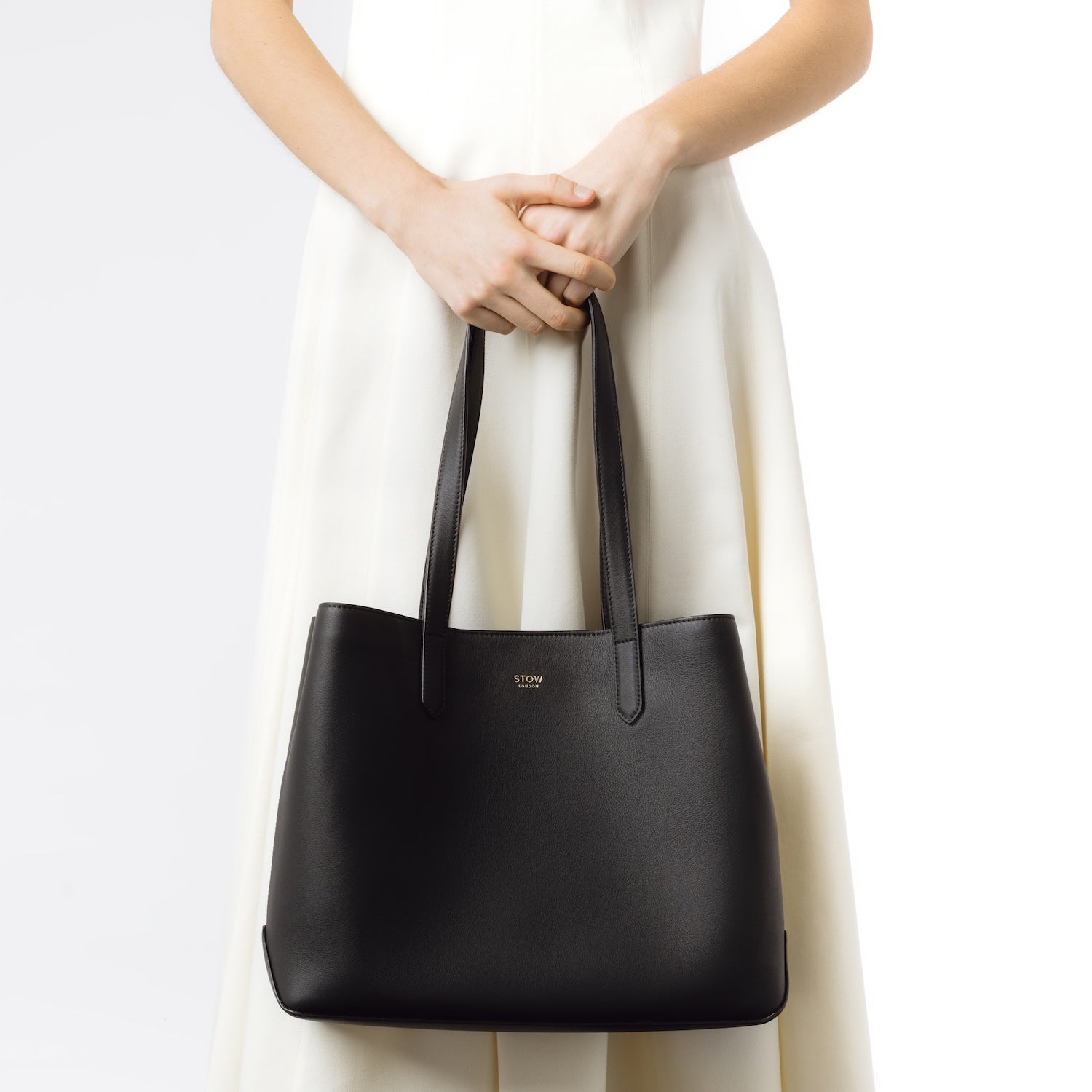 Model holding Stow London's Everyday Tote Bag in Black smooth leather on a light grey background featuring gold STOW logo
