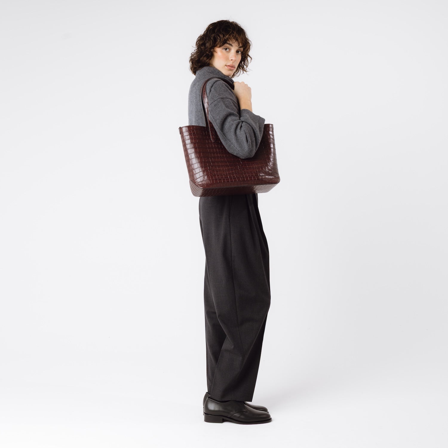 Model wearing Stow London's Everyday Tote Bag in Burgundy Embossed Croc leather on her shoulder featuring gold hardware on a white background