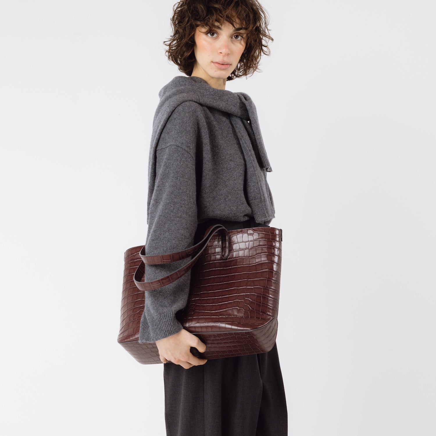 Model holding Stow London's Everyday Tote Bag in Burgundy Embossed Croc leather at her side featuring gold hardware on a white background