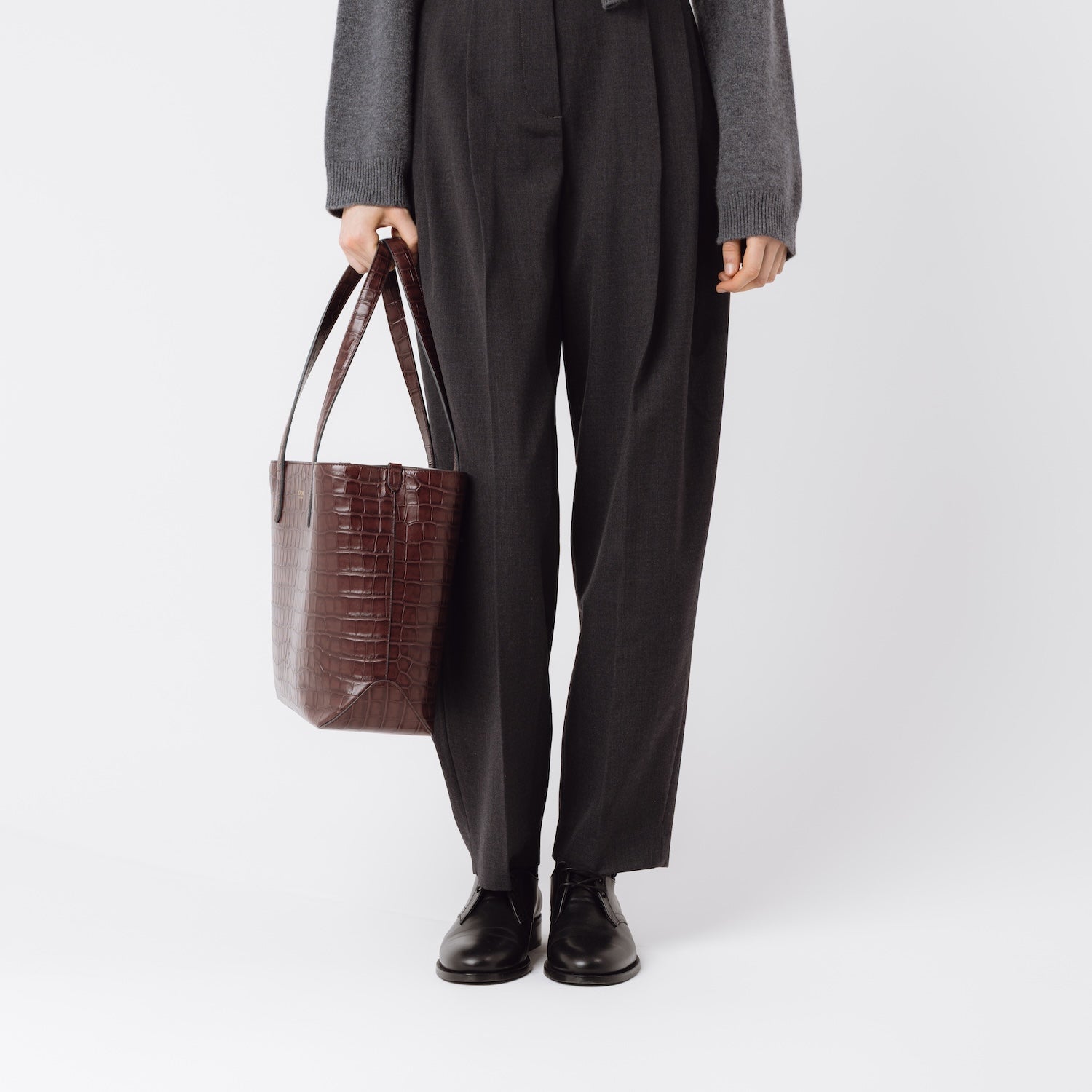 Model holding Stow London's Everyday Tote Bag in Burgundy Embossed Croc leather at her side on a white background