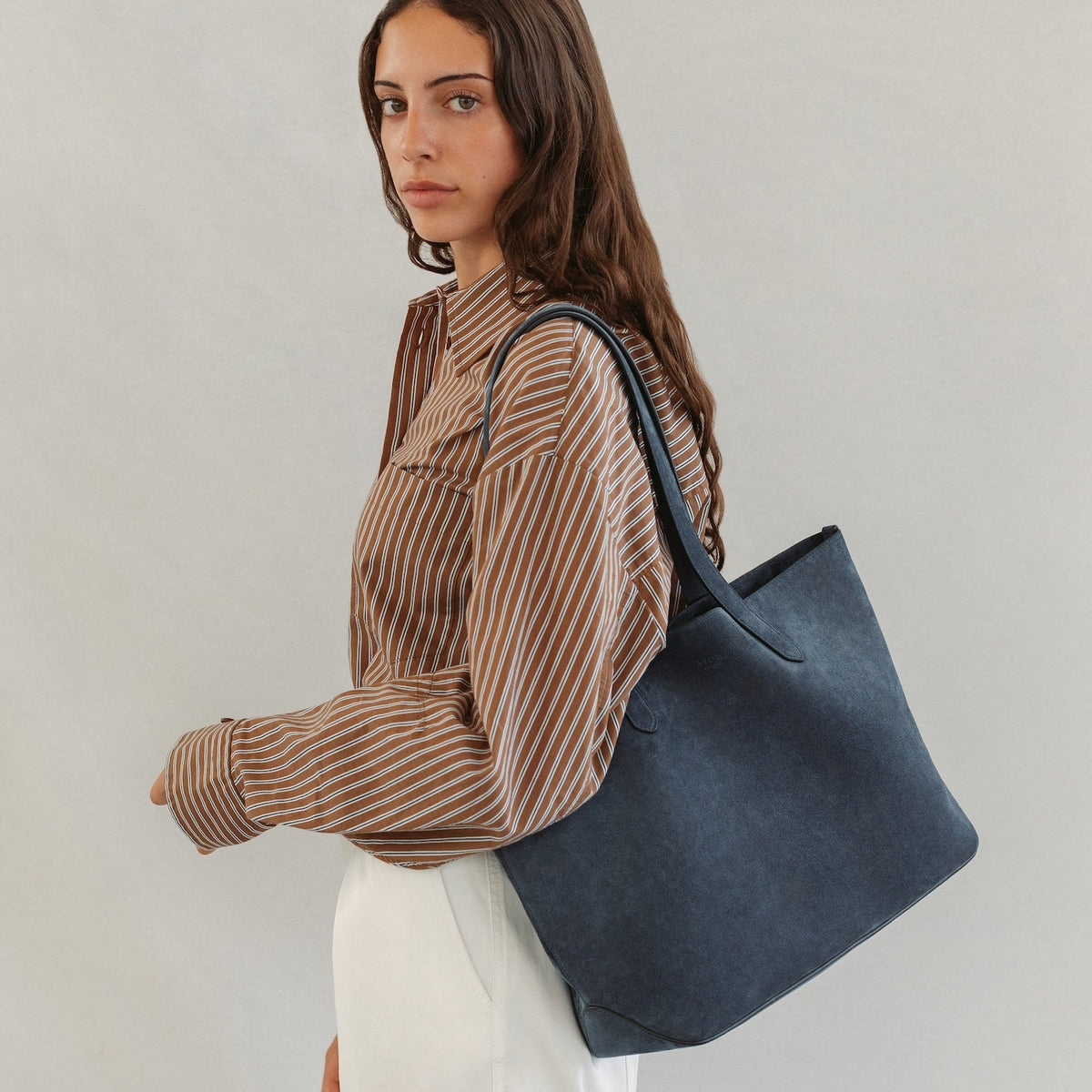 Model wearing STOW London's Everyday Tote bag in Navy suede on a light grey background featuring a small STOW London logo embossed on the front panel
