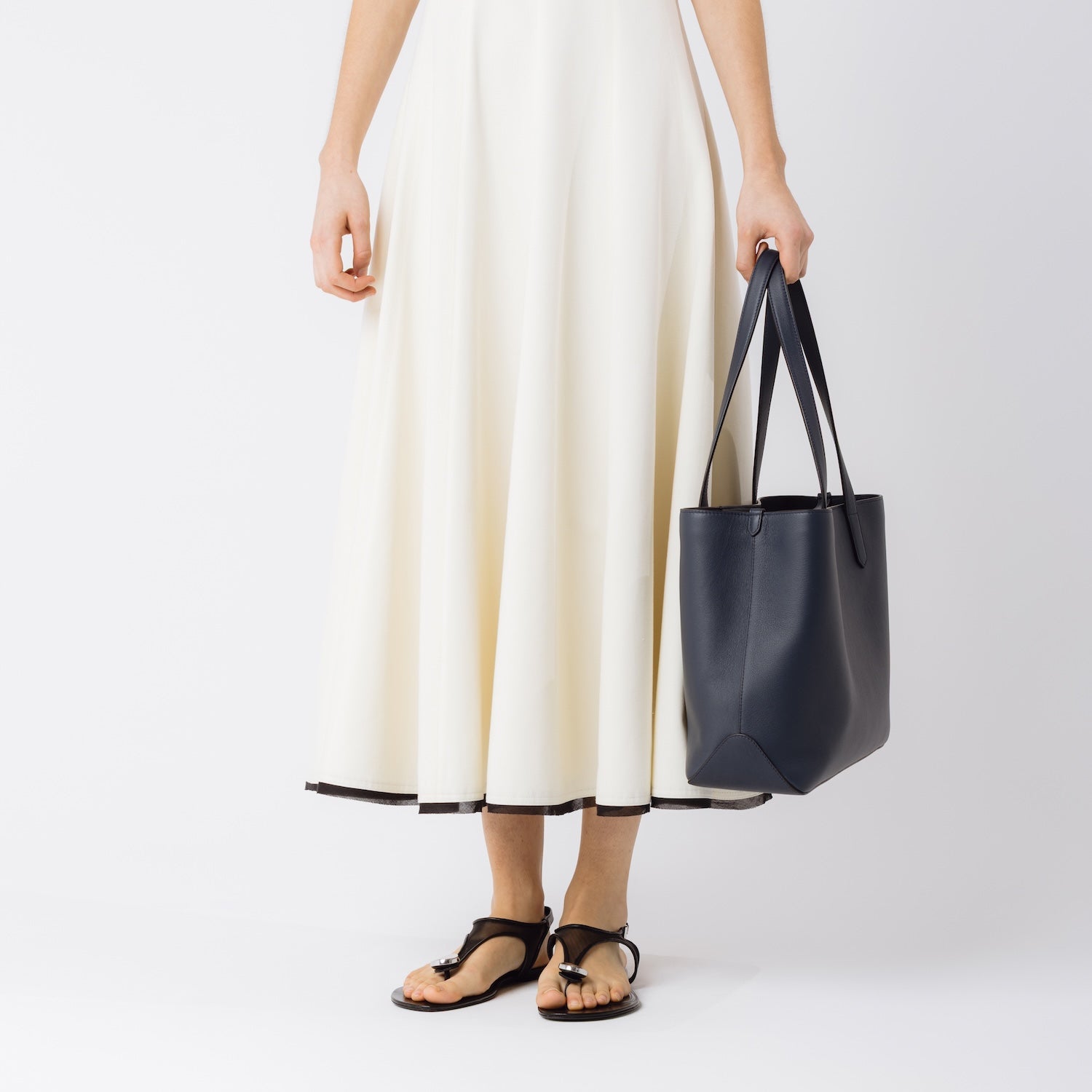 Model holding Stow London's Everyday Tote Bag in Navy smooth leather at her side on a white background