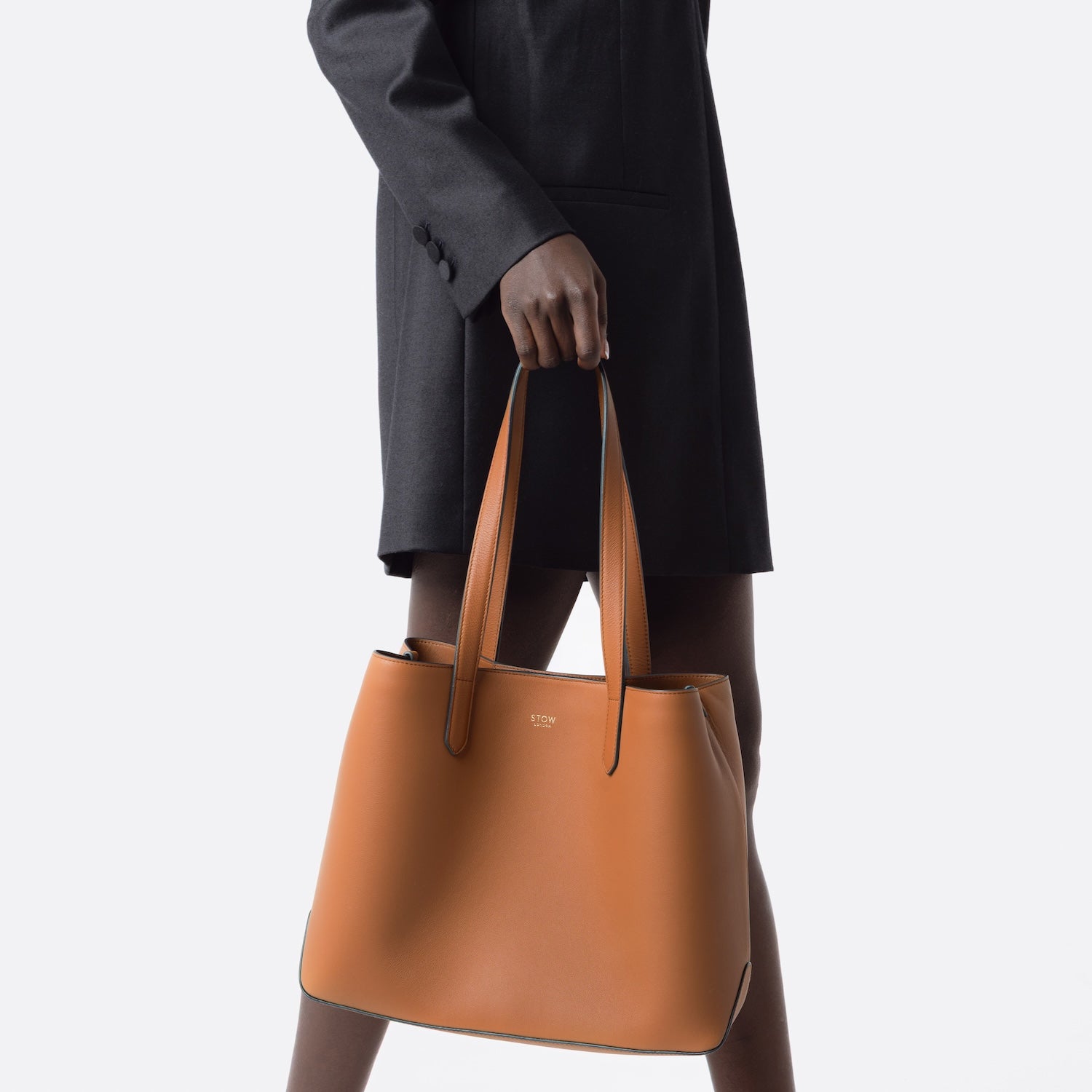 Model carrying STOW London's Everyday Tote bag in Earth Tan smooth leather by handles at her side on a white background