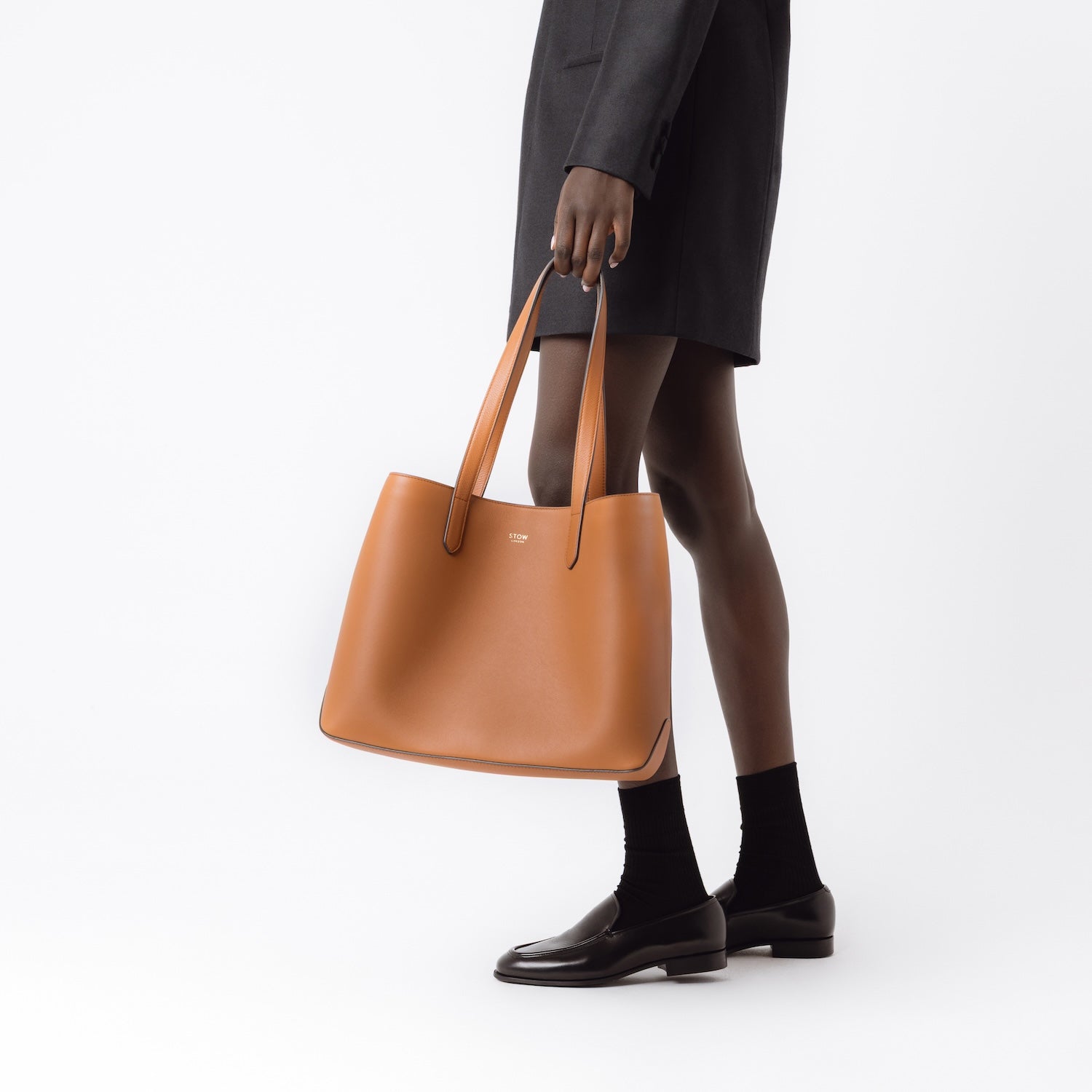 Model carrying STOW London's Everyday Tote bag in Earth Tan smooth leather by handles at her side on a white background