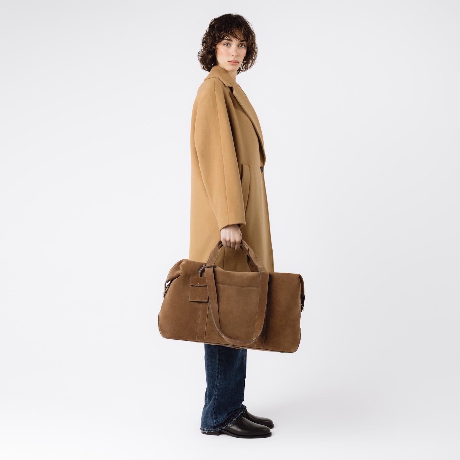 Model holding STOW London's Weekend bag in Chocolate suede leather at her side on a white background