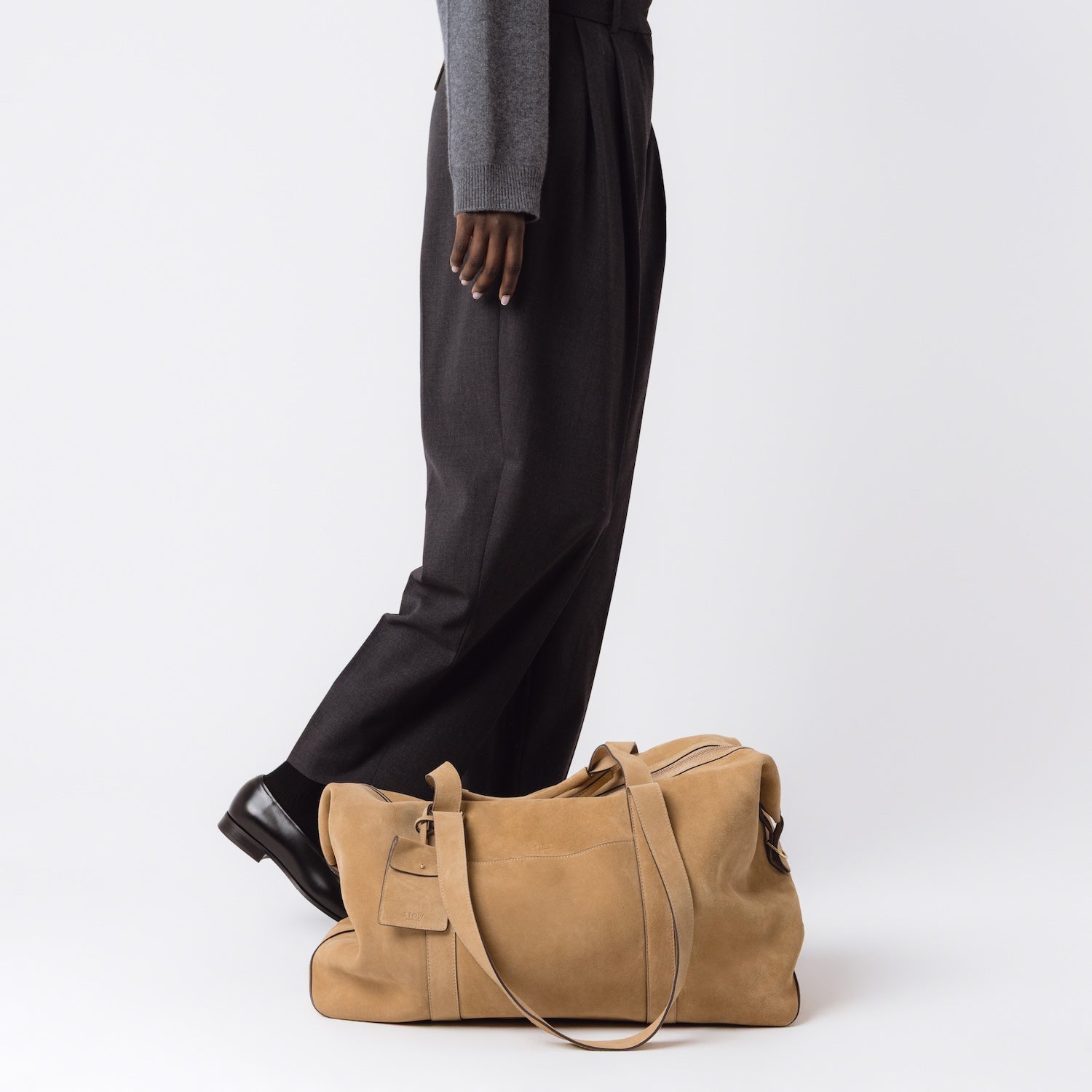 STOW London's Weekend bag in Soft Sand suede set down at model's feet on a light grey background