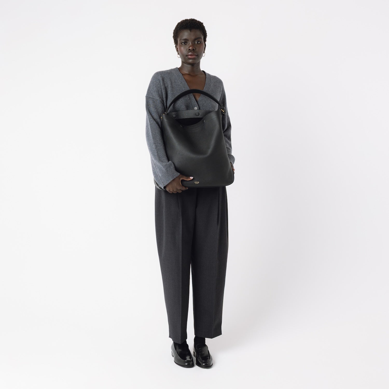 Model holding STOW London's Maxi Curve bag in Black pebbled leather featuring gold-coated stainless steel hardware on a light grey background