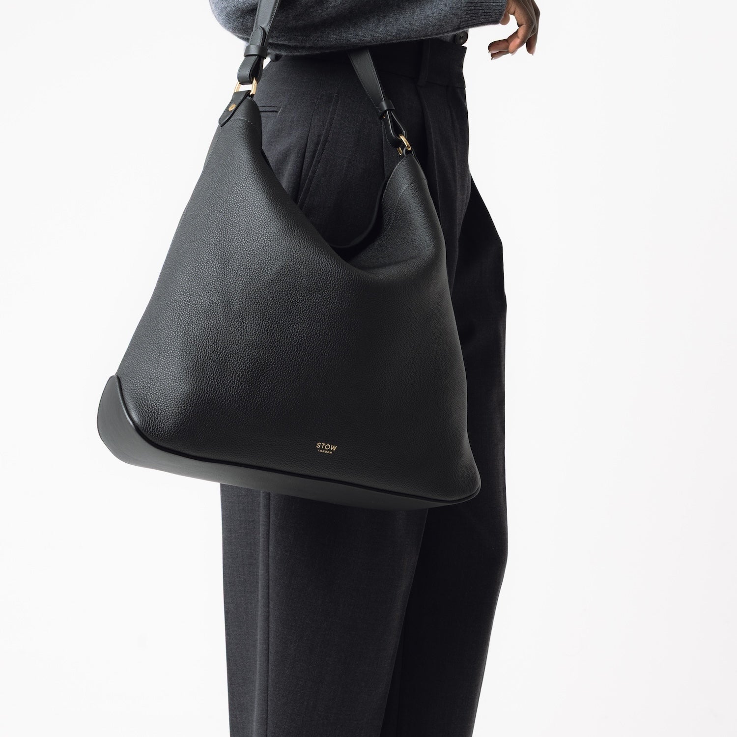 Model wearing STOW London's Maxi Curve bag in Black pebbled leather on her arm featuring gold-coated stainless steel hardware on a light grey background
