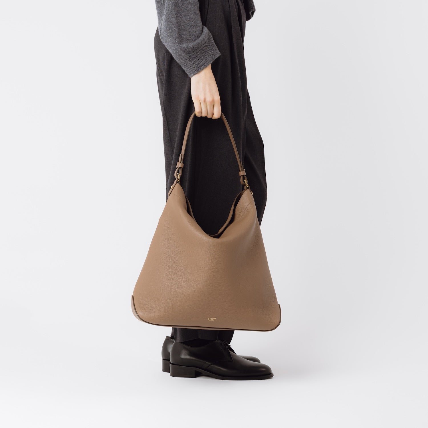 Model holding STOW London's Maxi Curve bag in Mocha pebbled leather at her side featuring gold-coated stainless steel hardware on a light grey background