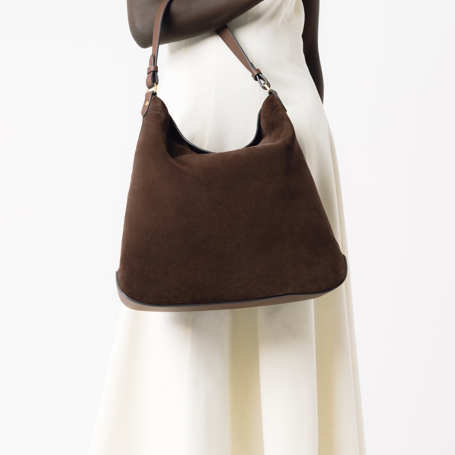 Model holding STOW London's Maxi Curve bag in Chocolate suede leather on her arm featuring gold-coated stainless steel hardware on a light grey background