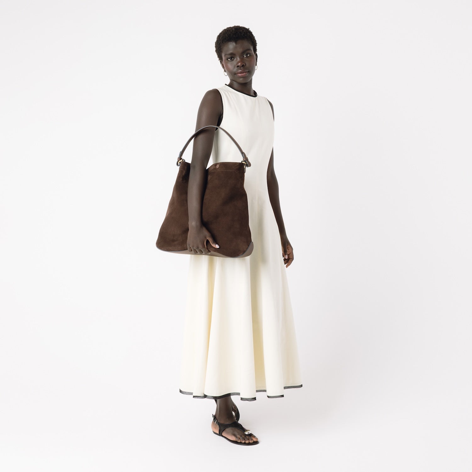 Model holding STOW London's Maxi Curve bag in Chocolate suede leather featuring gold-coated stainless steel hardware on a light grey background