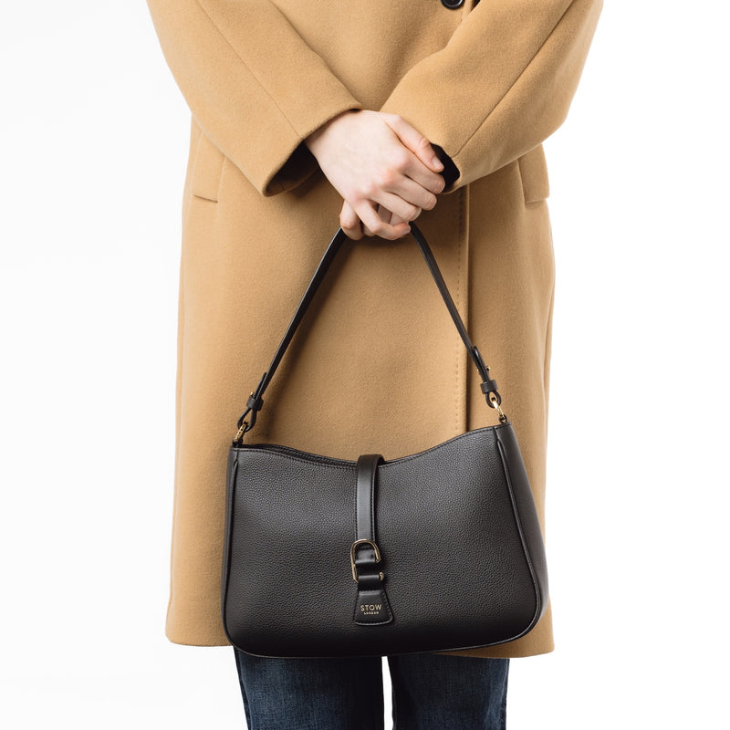 Model holding STOW London's Beth bag in Black pebbled and smooth leather at her front featuring gold-coated stainless steel hardware and clip on a white background