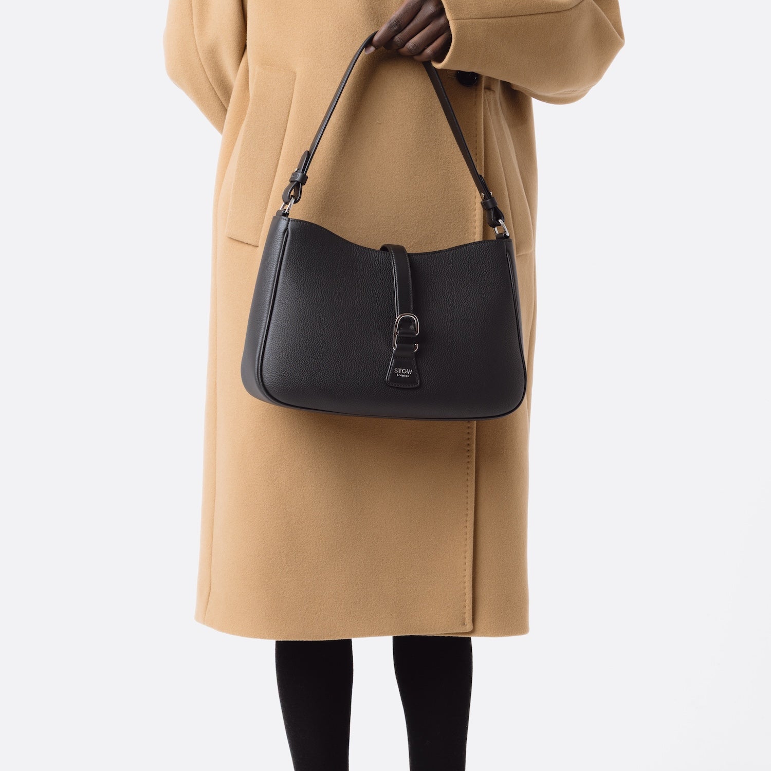 Model holding STOW London's Beth bag in Black pebbled and smooth leather at her front featuring silver stainless steel hardware and clip on a white background
