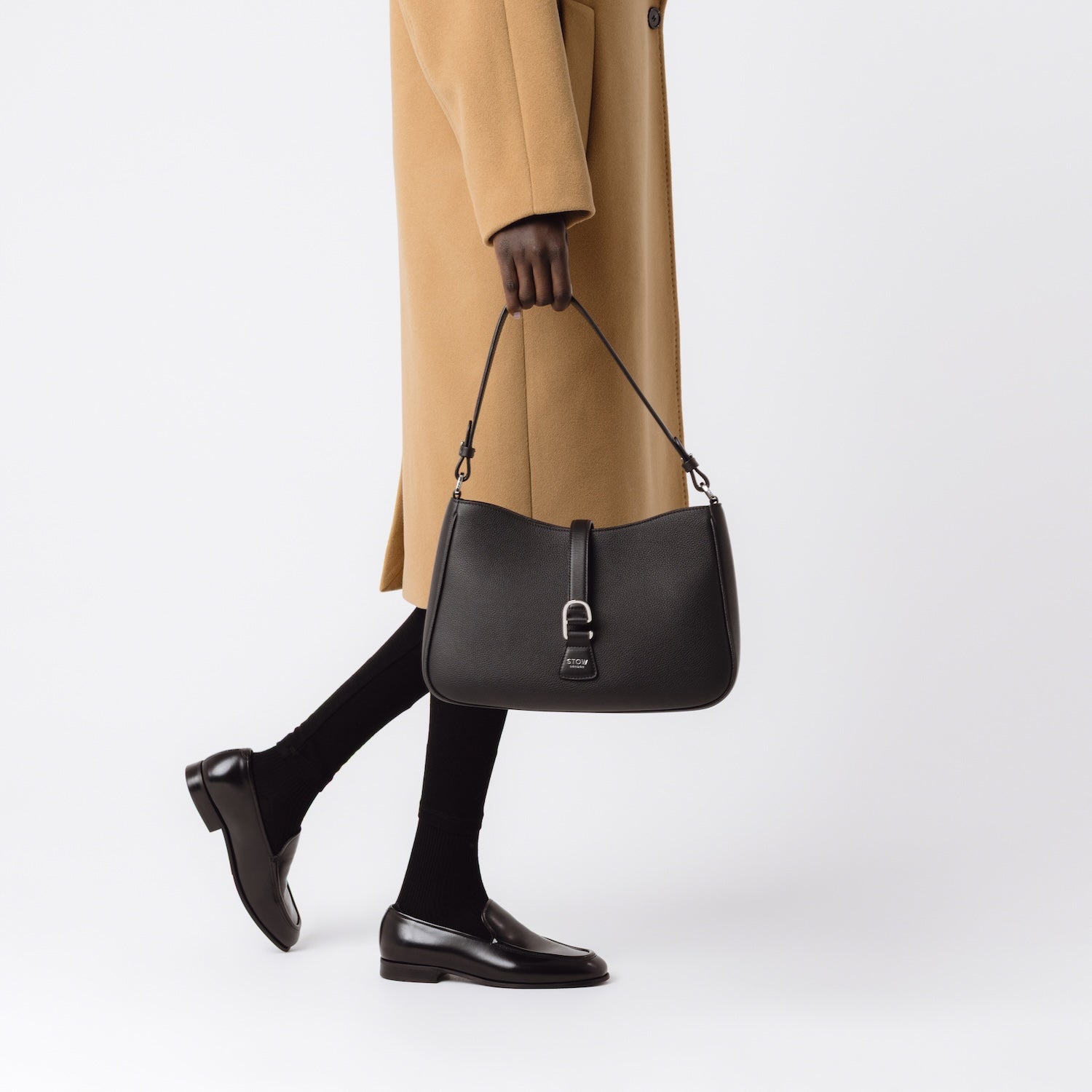 Model holding STOW London's Beth bag in Black pebbled and smooth leather at her side featuring silver stainless steel hardware and clip on a white background