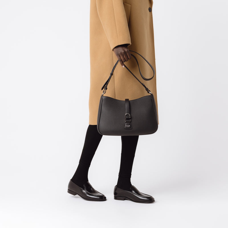 Model holding STOW London's Beth bag by its detachable crossbody strap in Black leather featuring silver stainless steel hardware on a white background