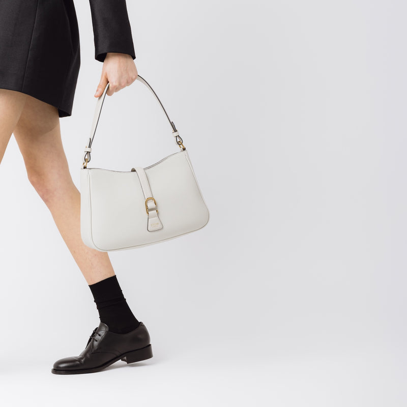 Model holding STOW London's Beth bag in Cloud Grey pebbled and smooth leather in her hand at her side featuring gold-coated stainless steel hardware and clip on a white background