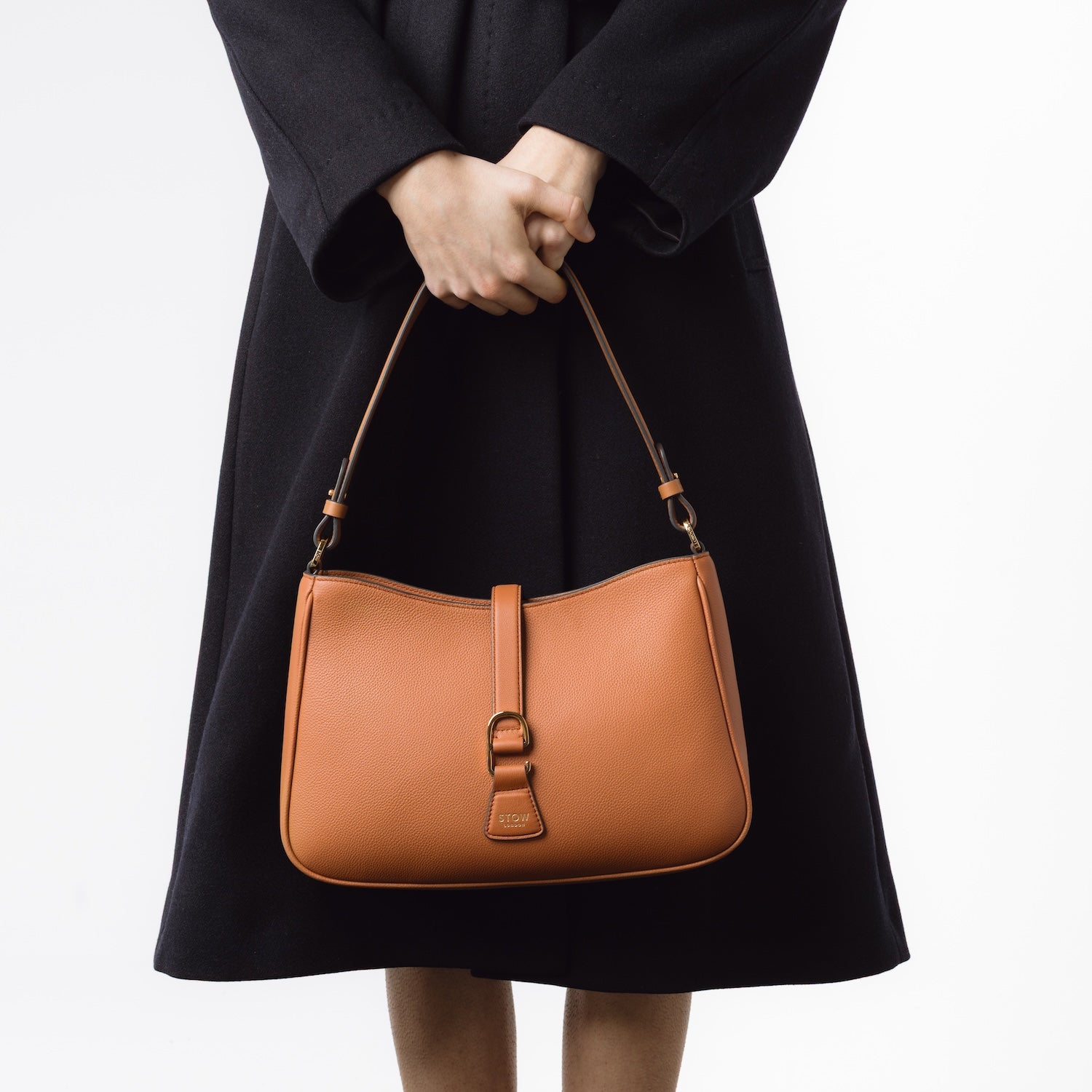 STOW London's Beth bag in Earth Tan leather held at front on model by its strap featuring gold-coated stainless steel hardware on a white background 