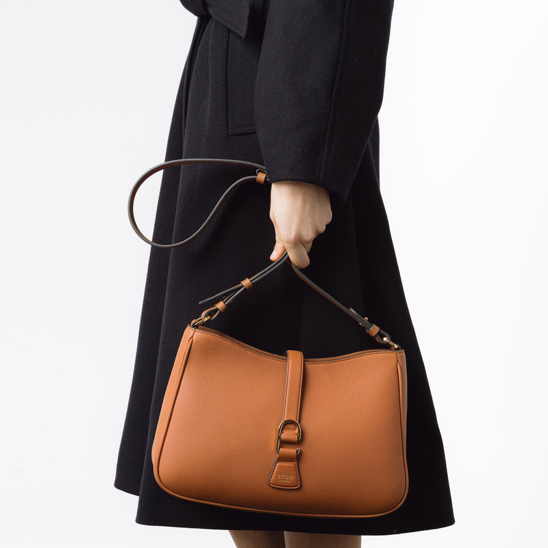 Model wearing STOW London's Earth Tan Beth bag by its crossbody strap at her side featuring gold-coated hardware on a white background