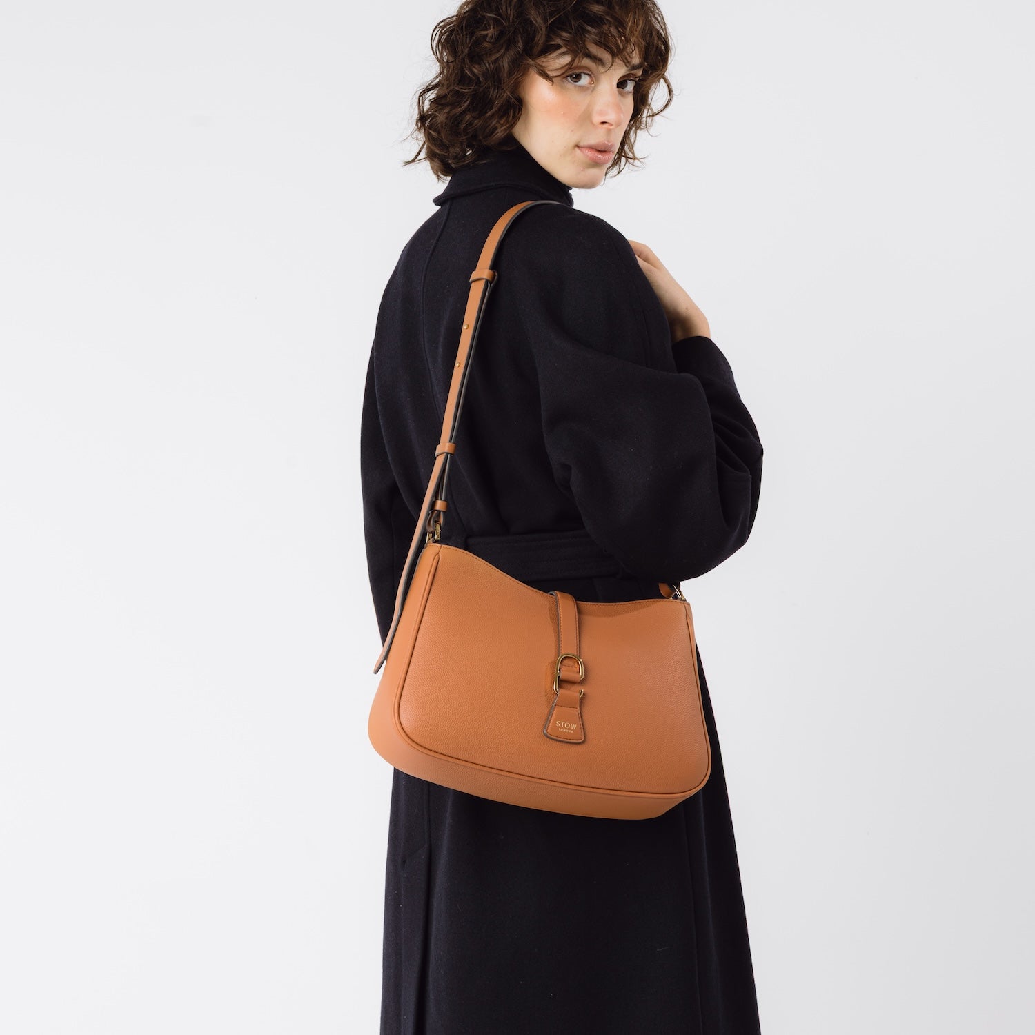 Model wearing STOW London's Earth Tan Beth bag on her shoulder with its detachable crossbody leather strap featuring gold-coated stainless steel hardware on a white background