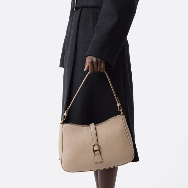Model holding STOW London's Beth bag in Warm Taupe pebbled and smooth leather at her side featuring gold-coated stainless steel hardware and clip on a white background
