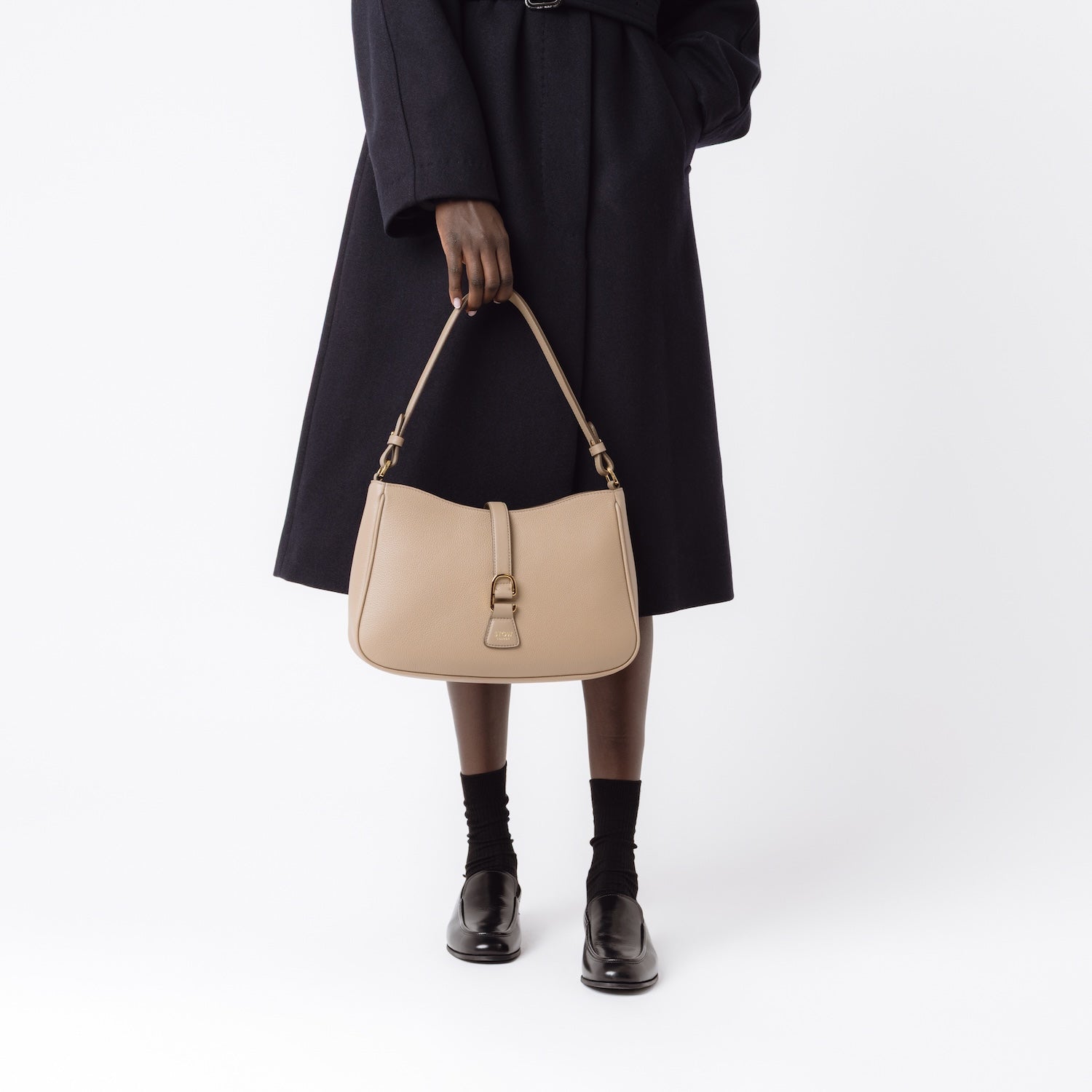 Model holding STOW London's Beth bag in Warm Taupe pebbled and smooth leather at her front featuring gold-coated stainless steel hardware and clip on a white background