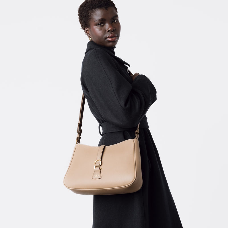 Model wearing STOW London's Beth bag by the detachable crossbody strap in Warm Taupe leather featuring gold stainless steel hardware on a white background