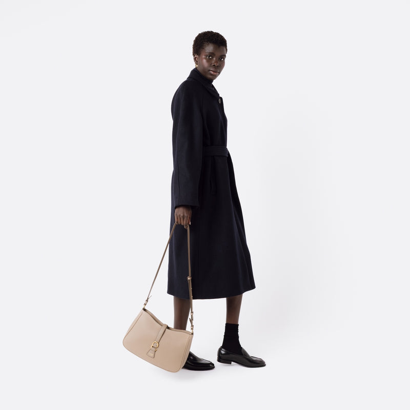 Model holding STOW London's Beth bag by the detachable crossbody strap in Warm Taupe leather at her side featuring gold stainless steel hardware on a white background