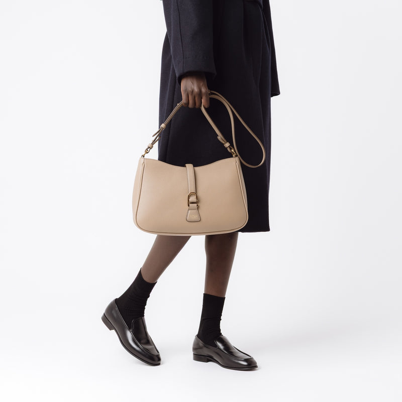 Model holding STOW London's Beth bag by the detachable crossbody strap in Warm Taupe leather featuring gold stainless steel hardware on a white background