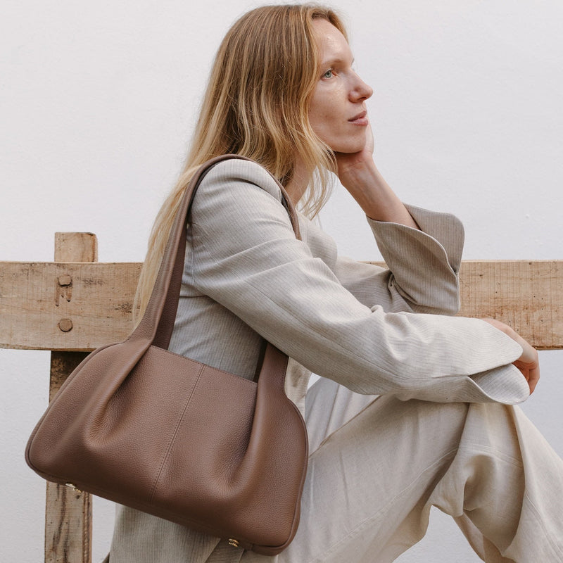 Model sat on a bench wearing STOW London's Bowling bag in Mocha pebbled leather on her shoulder, featuring gold-stainless steel hardware