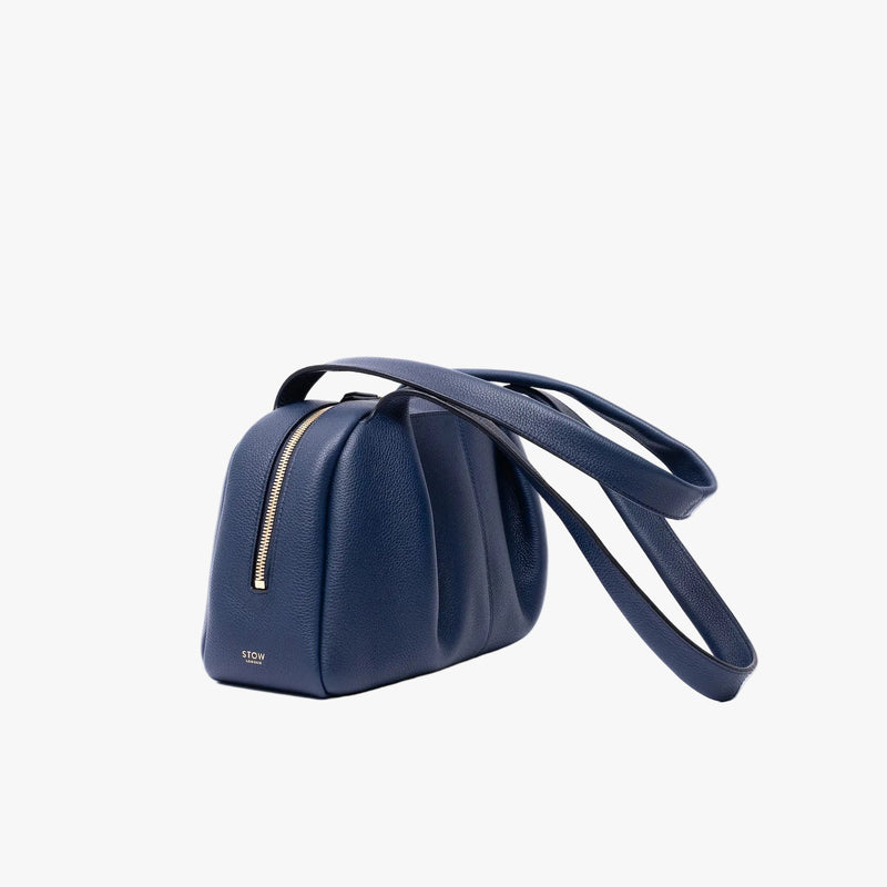 Off-axis view of STOW London's Bowling bag in Navy pebbled leather featuring the pleated front panel and gold-coated stainless steel hardware on a light grey background