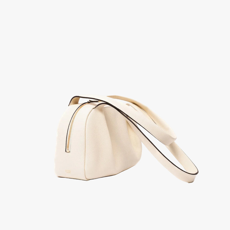 Off-axis view of STOW London's Bowling bag in Spring Moon pebbled leather featuring the pleated front panel and gold-coated stainless steel hardware on a light grey background