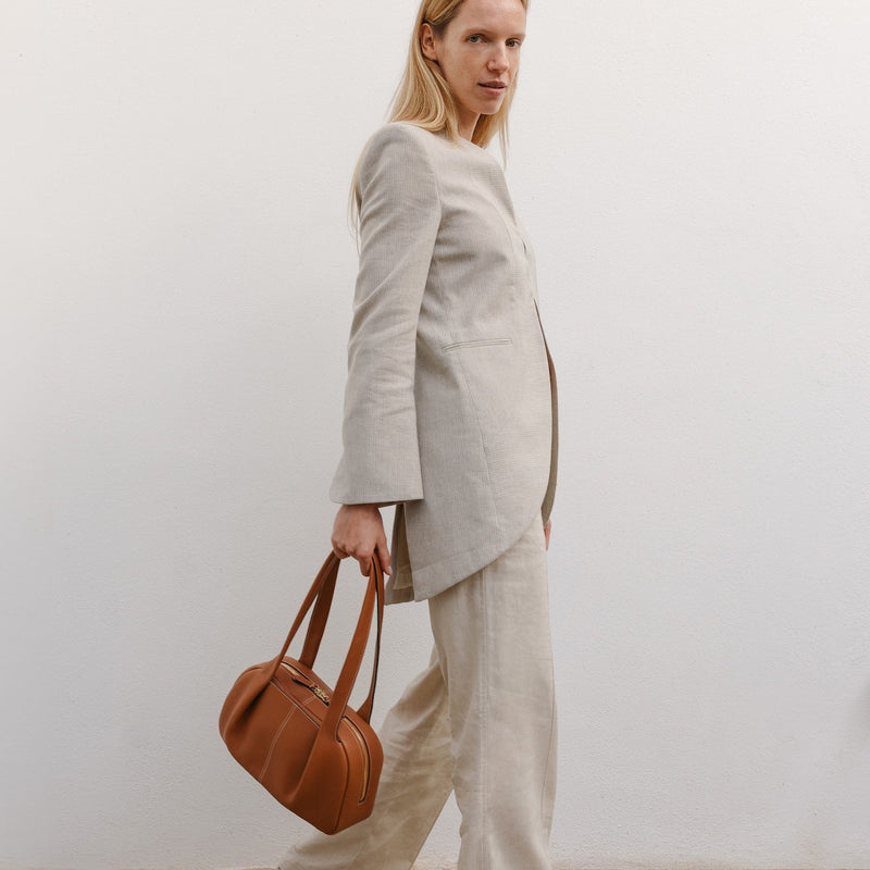 Model holding STOW London's Bowling bag in Earth Tan pebbled leather featuring the pleated front panel and gold-coated stainless steel hardware 