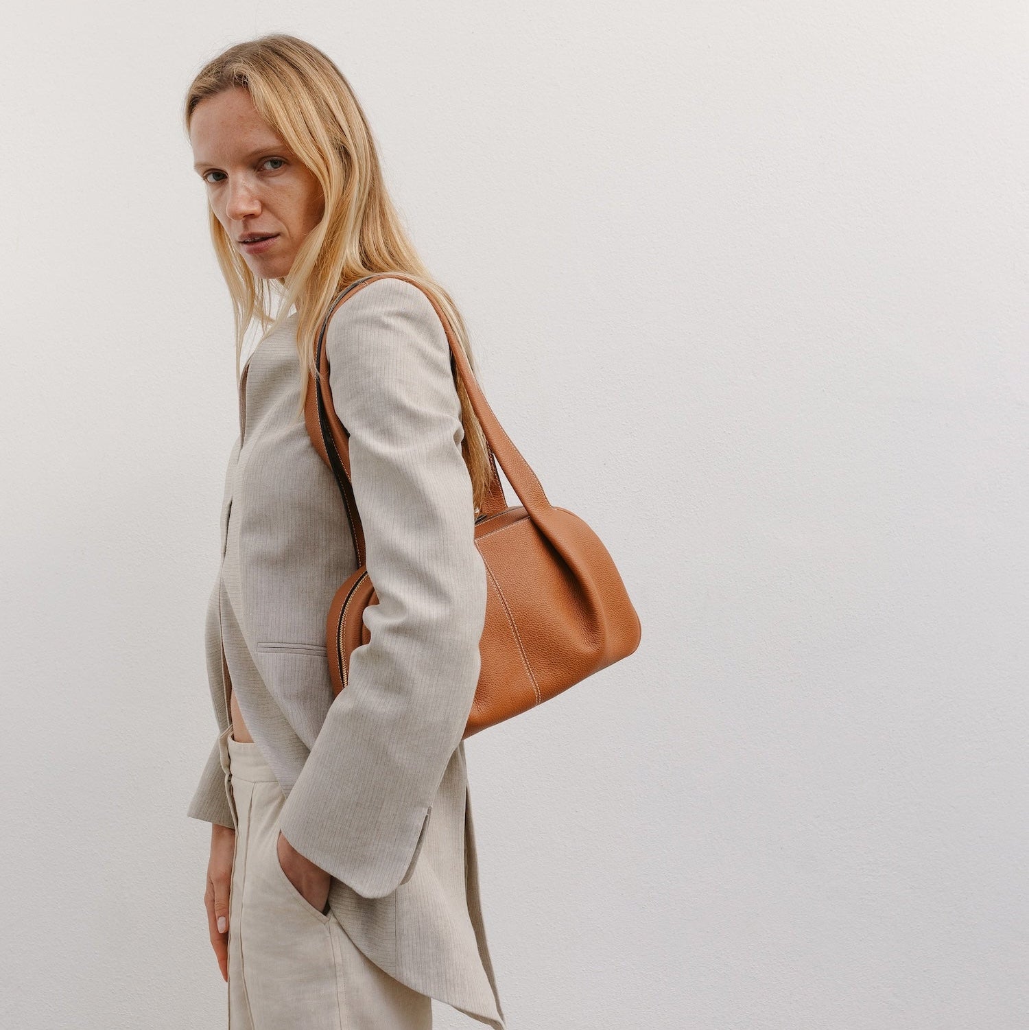 Model wearing STOW London's Bowling bag in Earth Tan pebbled leather on her shoulder featuring the pleated front panel and gold-coated stainless steel hardware 