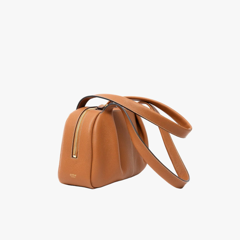 Off-axis view of STOW London's Bowling bag in Earth Tan pebbled leather featuring the pleated front panel and gold-coated stainless steel hardware on a light grey background