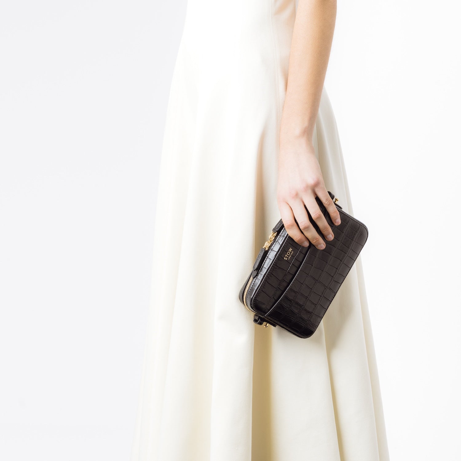 Model holding STOW London's Camera bag in Black Embossed Croc leather in her hand by her side featuring gold-coated stainless steel hardware on a white background