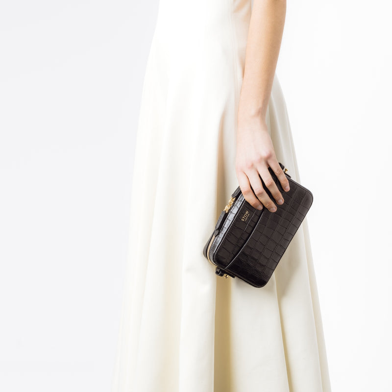Model holding STOW London's Camera bag in Black Embossed Croc leather in her hand by her side featuring gold-coated stainless steel hardware on a white background