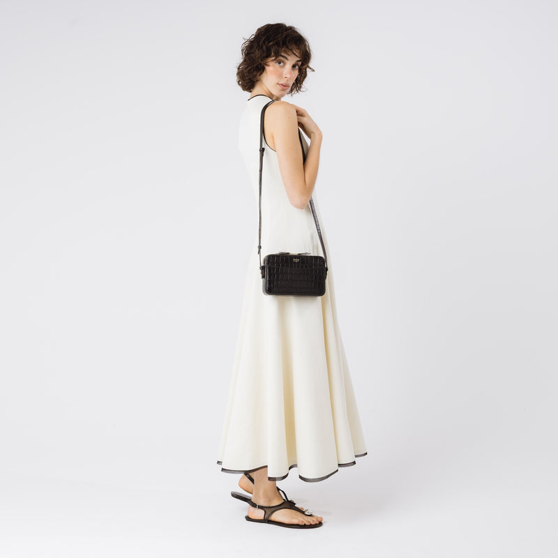 Model wearing STOW London's Camera bag in Black Embossed Croc leather on her shoulder with adjustable strap featuring gold-coated stainless steel hardware on a white background