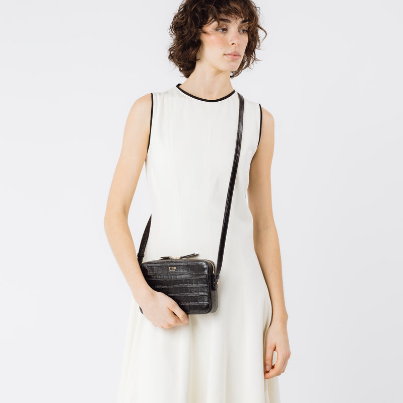Model wearing STOW London's Camera bag in Black Embossed Croc leather crossbody with adjustable strap featuring gold-coated stainless steel hardware on a white background