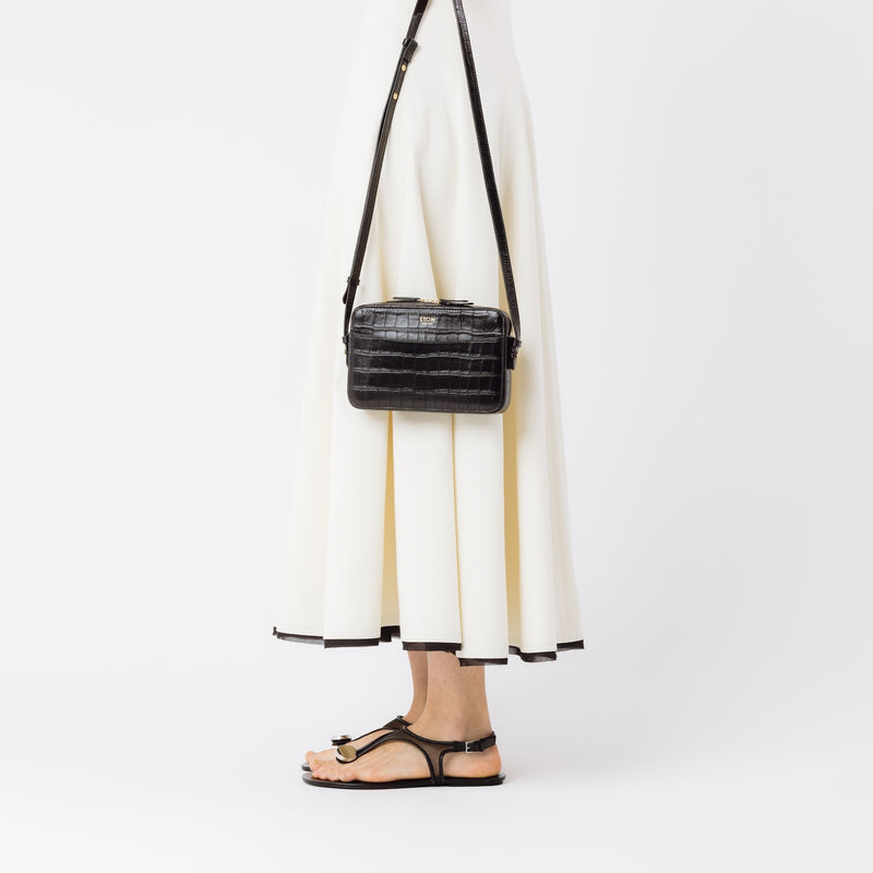 Model holding STOW London's Camera bag in Black Embossed Croc leather with detachable crossbody strap featuring gold-coated stainless steel hardware on a white background