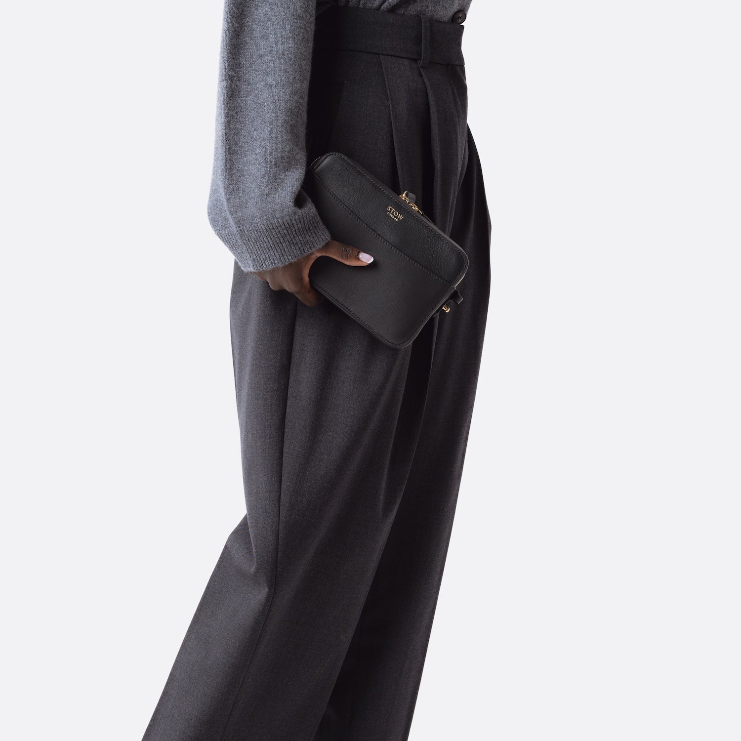 Model holding STOW London's Camera bag in Black smooth leather in hand at her side featuring gold-coated stainless steel hardware on a white background