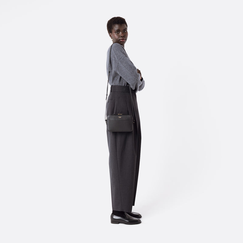 Model wearing STOW London's Camera bag in Black smooth leather on her shoulder with adjustable strap featuring gold-coated stainless steel hardware on a white background