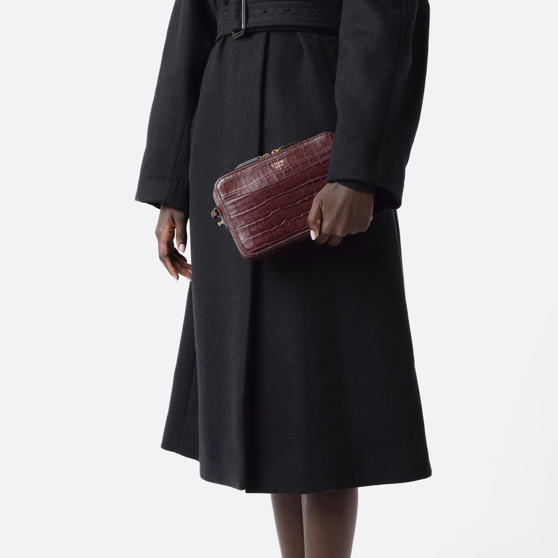 Model holding STOW London's Camera bag in Burgundy Embossed Croc leather in hand by her side featuring gold-coated stainless steel hardware on a white background