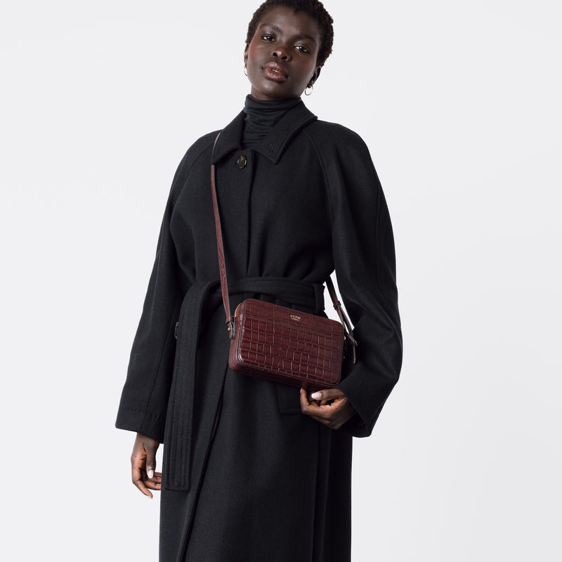 Model wearing STOW London's Camera bag in Burgundy Embossed Croc leather crossbody with adjustable strap featuring gold-coated stainless steel hardware on a white background