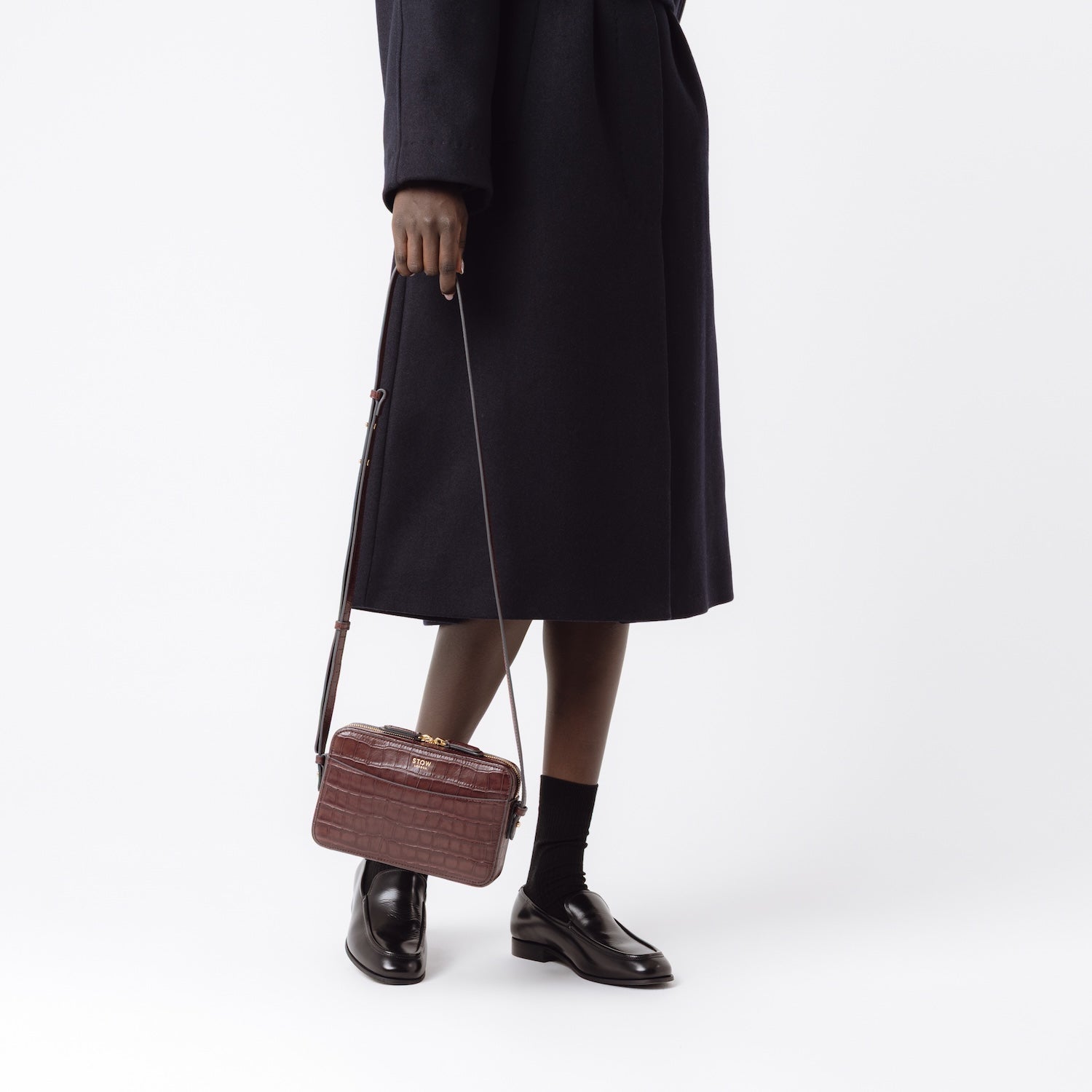 Model holding STOW London's Camera bag in Burgundy Embossed Croc leather by its crossbody strap at her side featuring gold-coated stainless steel hardware on a white background