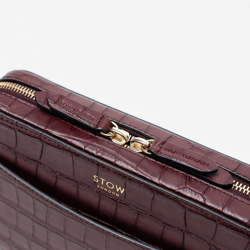 Close up of STOW London's Camera bag in Burgundy Embossed Croc leather featuring gold-coated stainless steel hardware and brand engraved pull tabs on a white background