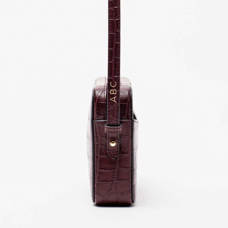 Side view of STOW London's Camera bag in Burgundy Embossed Croc leather showcasing the personalisation positioning and featuring gold-coated stainless steel hardware on a white background