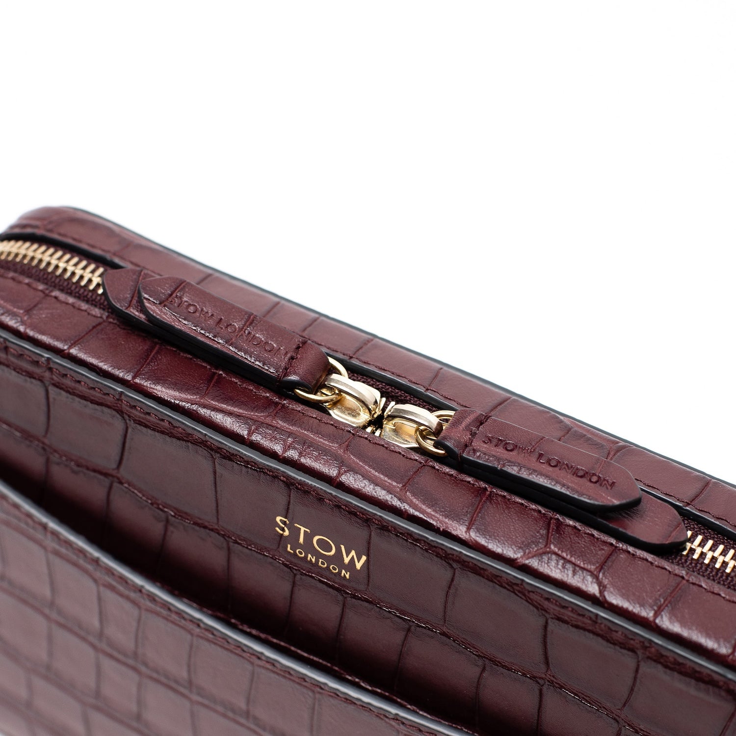 Close up view of STOW London's Camera bag in Burgundy Embossed Croc leather featuring gold-coated stainless steel hardware and brand engraved pull tabs on a white background