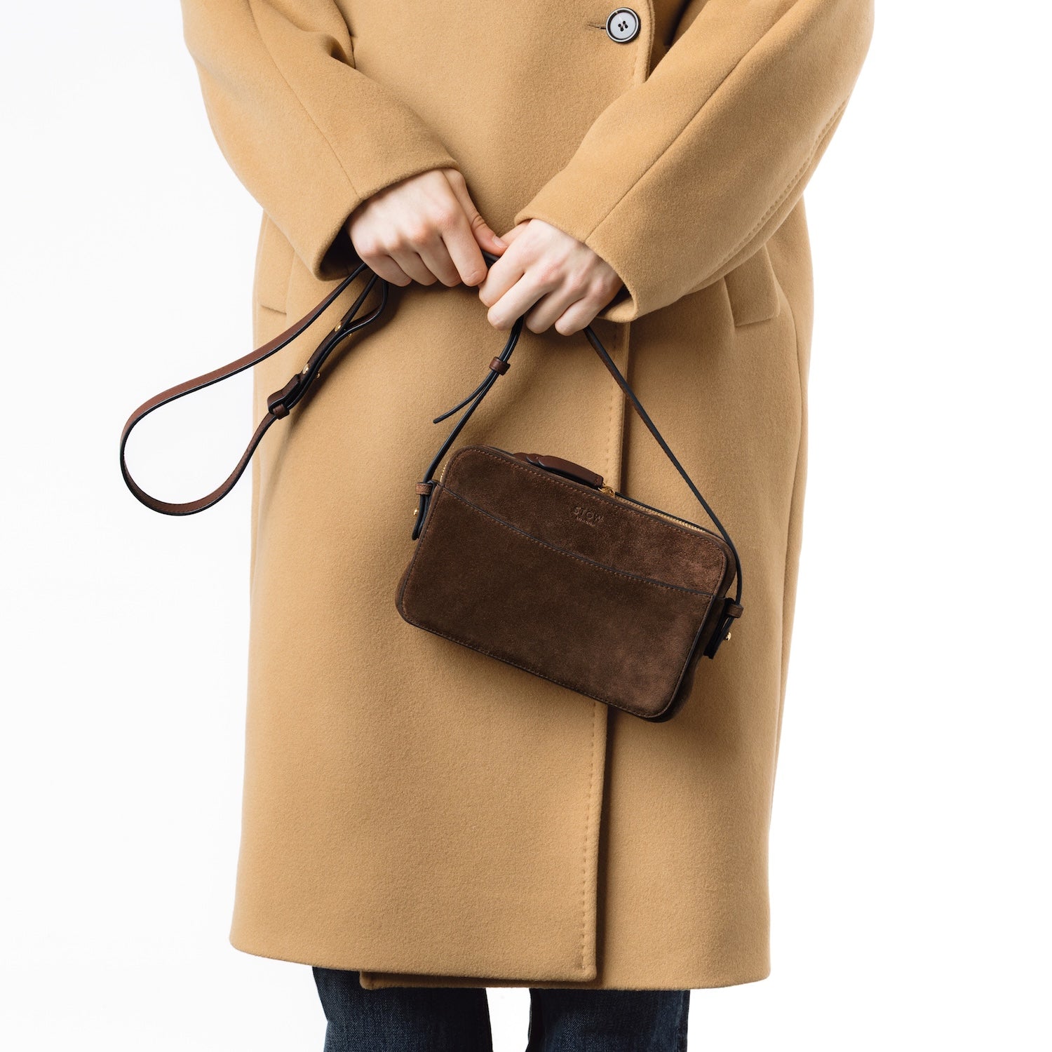 Model holding STOW London's Camera bag in Chocolate Suede leather by its detachable strap at her front featuring gold-coated stainless steel hardware on a white background